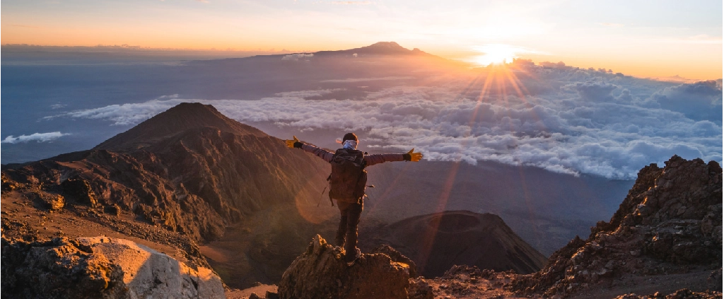 A hiker with arms wide open stands on a rocky mountain peak looking at a bright sunrise over a blanket of clouds, with a large mountain peak visible in the far distance.
