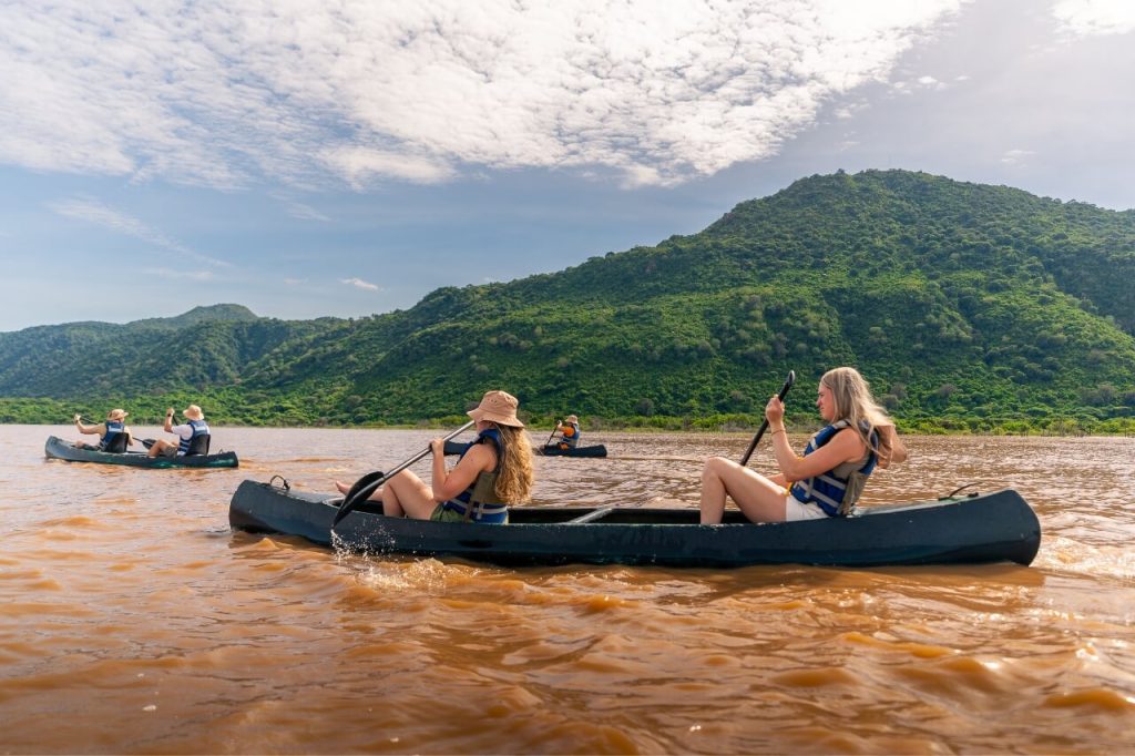  Four people in two black canoes paddle across wide, brown water with high, bright green mountains rising in the background under a cloudy blue sky.