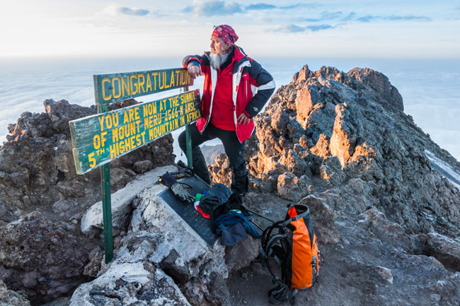 A male hiker in a red jacket and bandana leaning against a rustic wooden sign marking the summit of Mount Meru above a thick blanket of white clouds.
