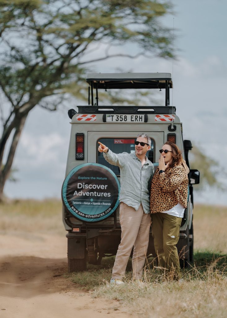 A man points out into the distance while standing beside a woman with an amazed expression next to a safari vehicle with a pop-up roof on a dirt path in the Serengeti.