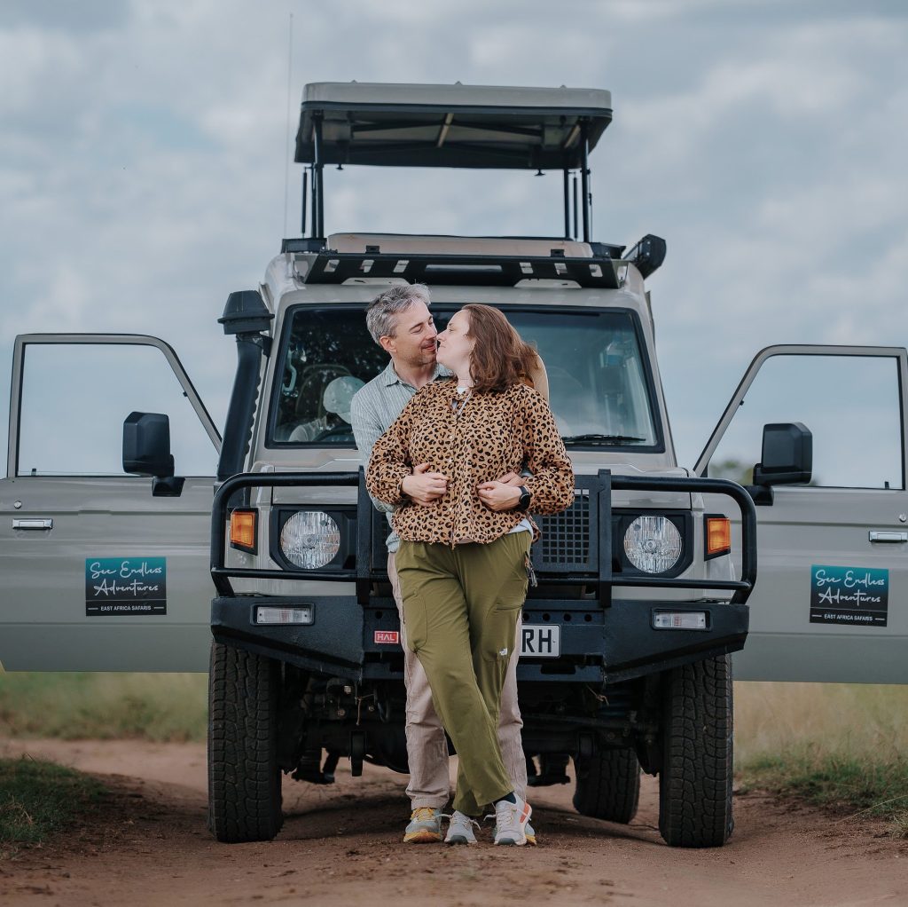  A man and a woman in safari clothing embrace and kiss while leaning against the front bumper of a 4x4 land cruiser on a dirt track under an overcast sky