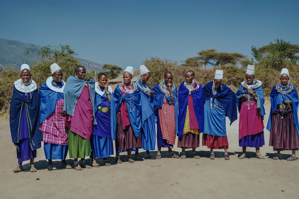 A line of Maasai women standing outside in a row, wearing bright blue and pink shΓΊkΓ cloaks, intricate beaded collars, and traditional white headpieces against a clear blue sky.