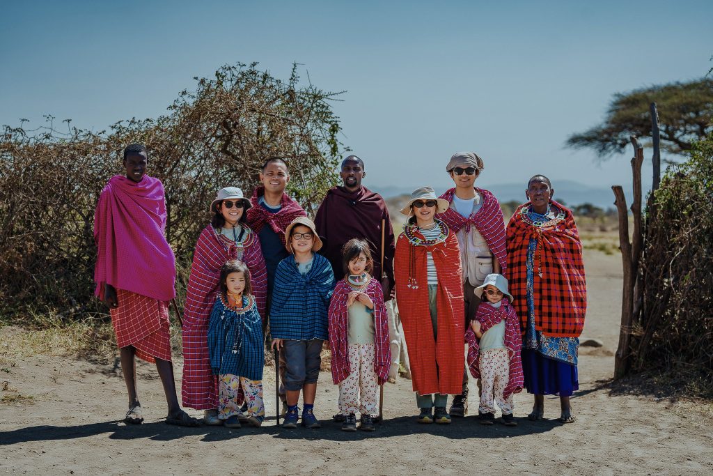 A group of international tourists, including young children, wearing Maasai shukas while posing alongside Maasai hosts near a village enclosure.