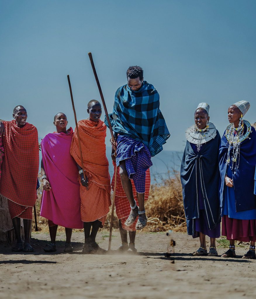 A Maasai man in a blue and black checked cloth jumps high off the ground holding a long stick, while men and women in colorful clothes and large beaded neck collars stand behind him singing.
