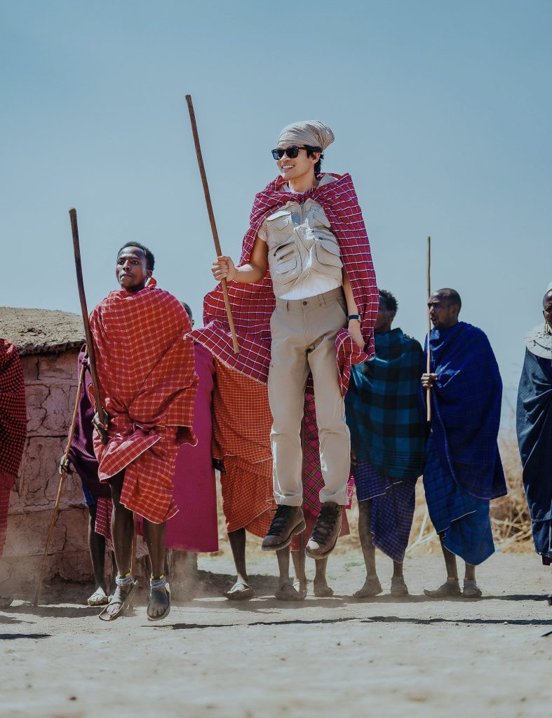 A male tourist wearing sunglasses, a utility vest, and a red plaid wrap leaping high into the air alongside Maasai men holding walking sticks during a traditional jumping dance.