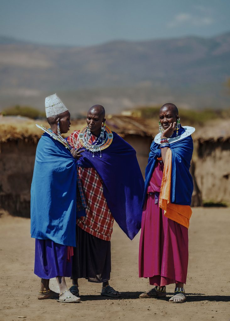 Three Maasai women wearing blue and purple wraps and large, flat, circular white beaded necklaces stand together talking outside on a sunny day.