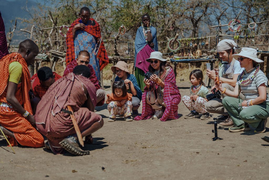 A group of foreign tourists, including young children and adults with cameras, sit in a circle on the ground watching Maasai men in traditional red clothing start a fire, with Maasai women standing behind them near a jewelry display.
