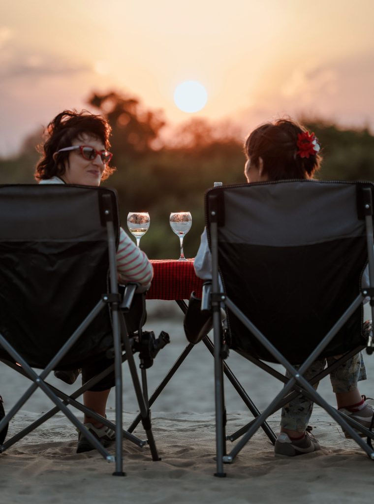 Two people sit facing each other in camping chairs on a sandy patch, with two glasses of water or wine on a small table between them as the sun sets in the background.