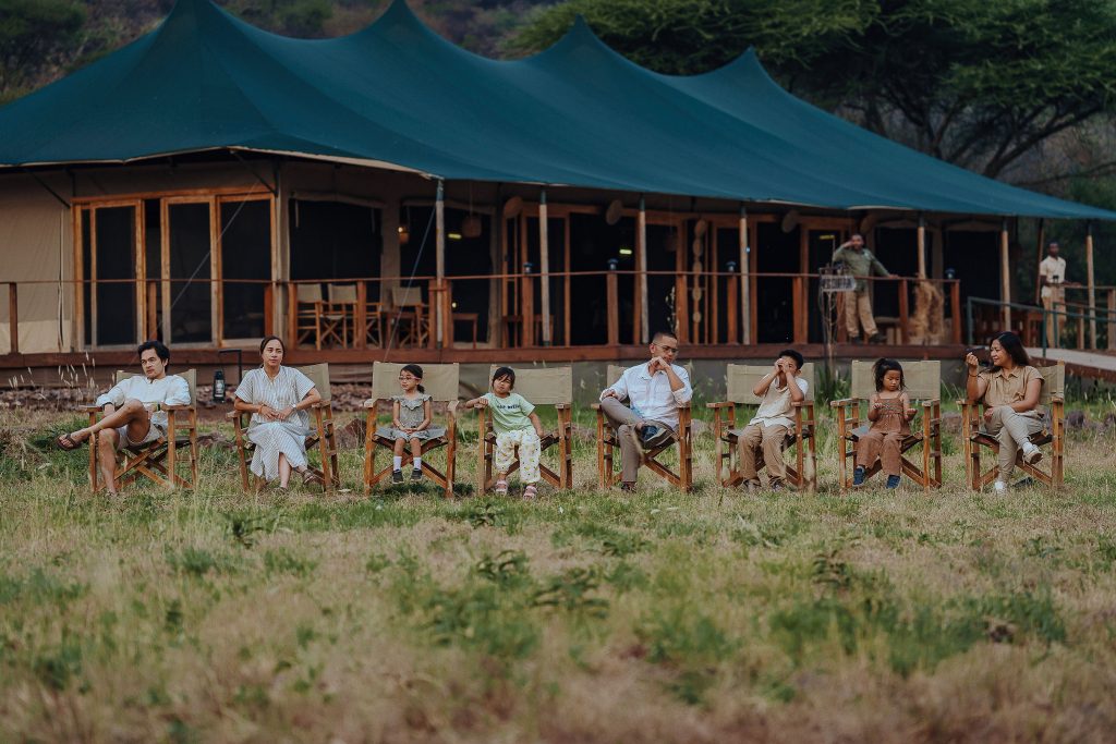 A large family group of eight people sitting side-by-side in canvas safari chairs in front of a massive dark green luxury safari tent structure on the grassy plains.
