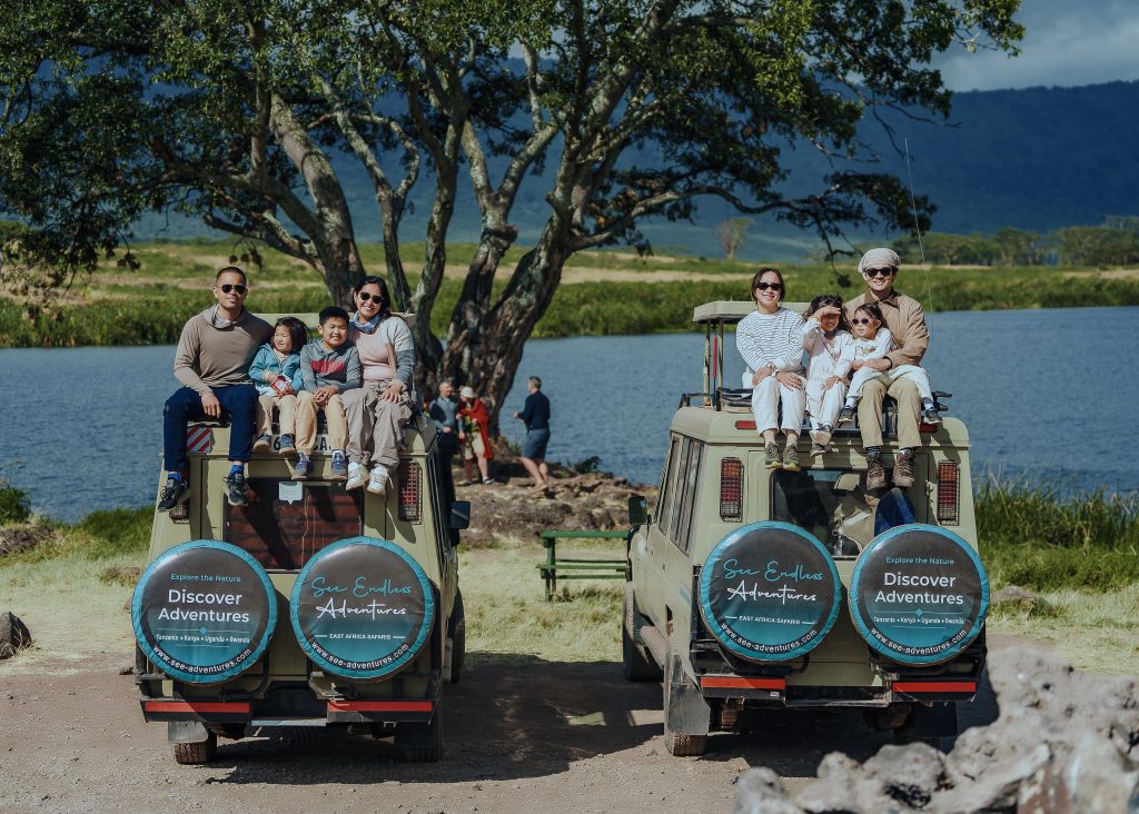 Two family groups of tourists sit on the roofs of two parked safari vehicles next to a large blue lake, accompanied by their drivers standing beside the spare tire covers.
