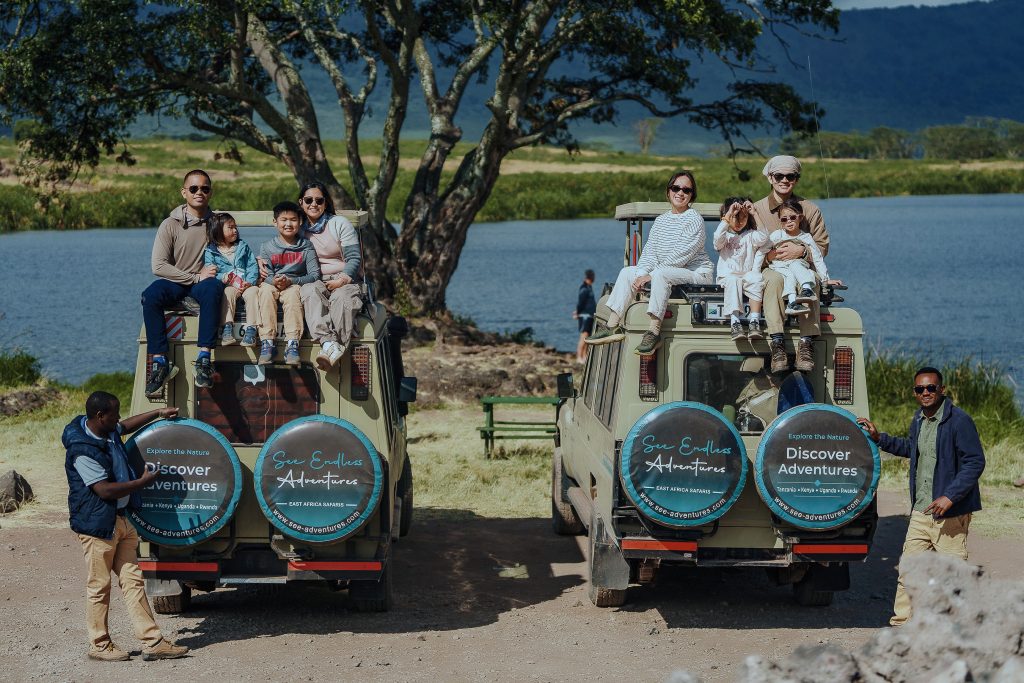 wo matching safari vehicles are parked side by side near a lake, with two families sitting on the roofs and their local guides standing nearby.