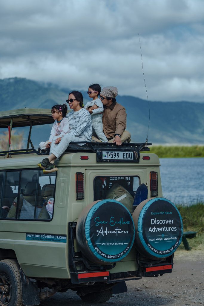A family sits safely on top of a safari vehicle looking out across a wide valley with misty green mountains under a cloudy sky.