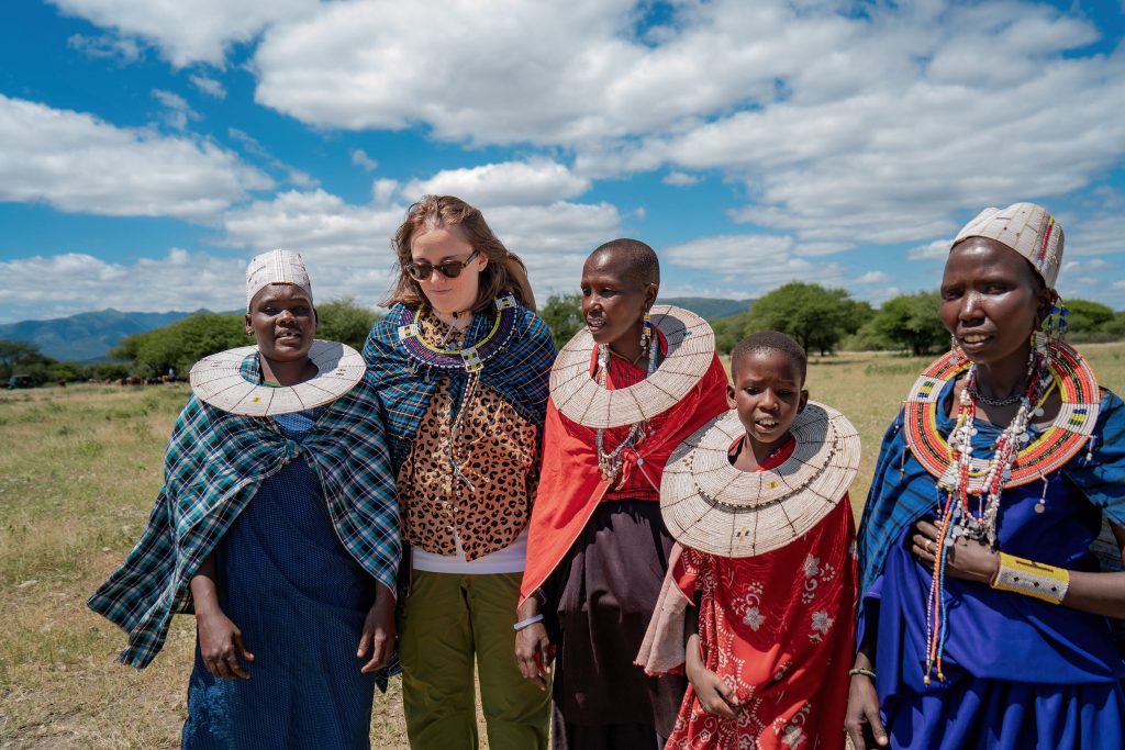 A group of five Maasai women and a young boy, along with a blonde female tourist wearing sunglasses, stand together in a sunny open field wearing traditional large beaded neck collars and colorful shúkà wraps.