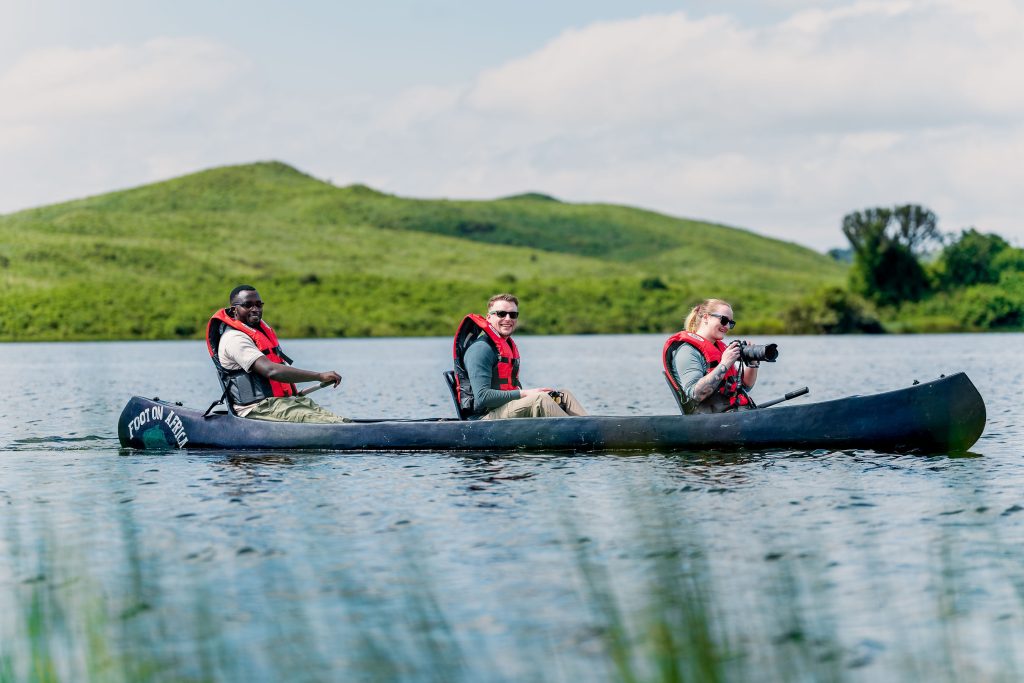 A side view of three people in a long black canoe on a calm lake, with a woman at the front holding a camera with a large lens and looking out toward rolling green hills.
