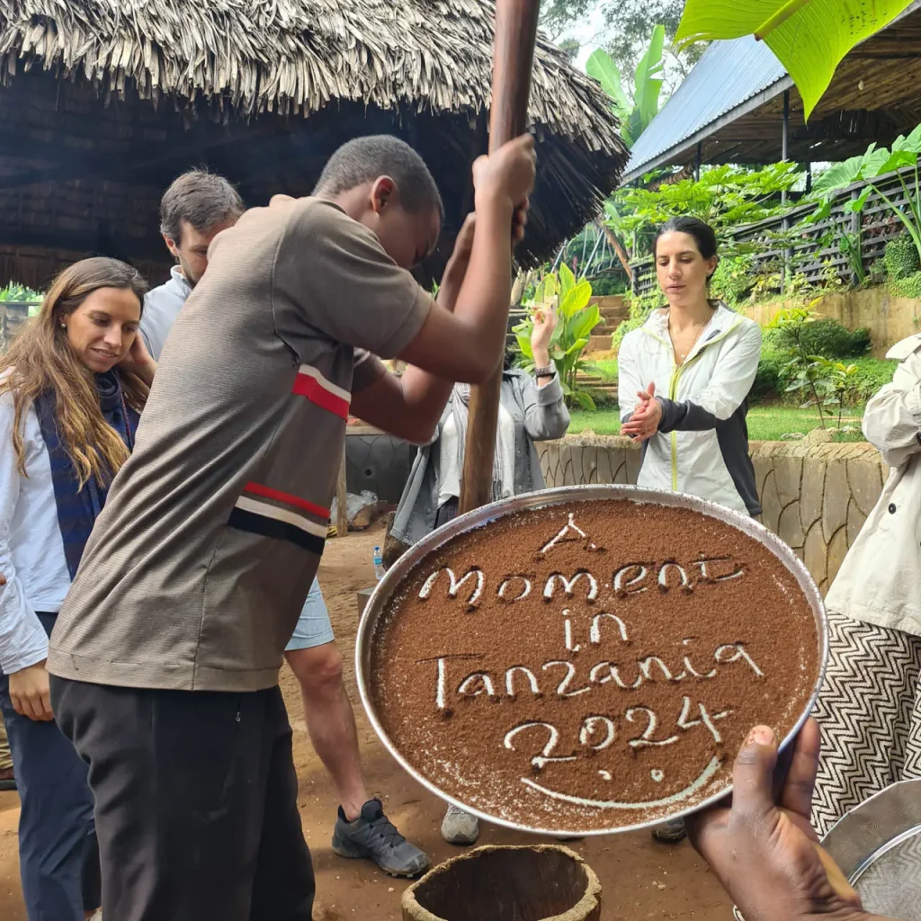 A young Tanzanian man grinding coffee beans using a large wooden mortar and pestle, while a shallow metal tray in the foreground displays ground coffee spelling out "A moment in Tanzania 2024".