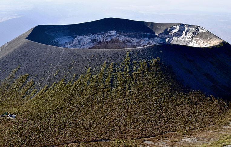 A high-angle aerial view of the massive, circular main crater rim of Ol Doinyo Lengai, with its steep outer slopes covered in green sparse vegetation and a tiny cluster of expedition tents visible at the lower left base.