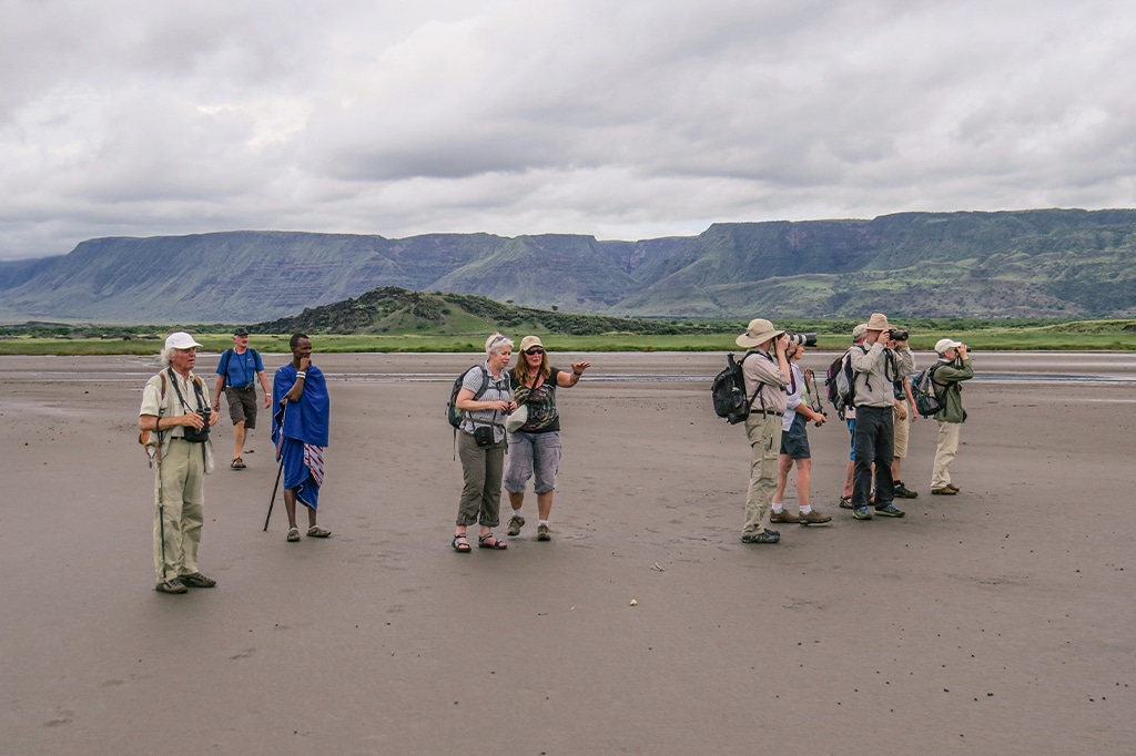 A group of foreign tourists wearing sun hats and backpacks stand on a vast dry mudflat, looking through binoculars and pointing toward distant escarpments with a local guide dressed in blue.