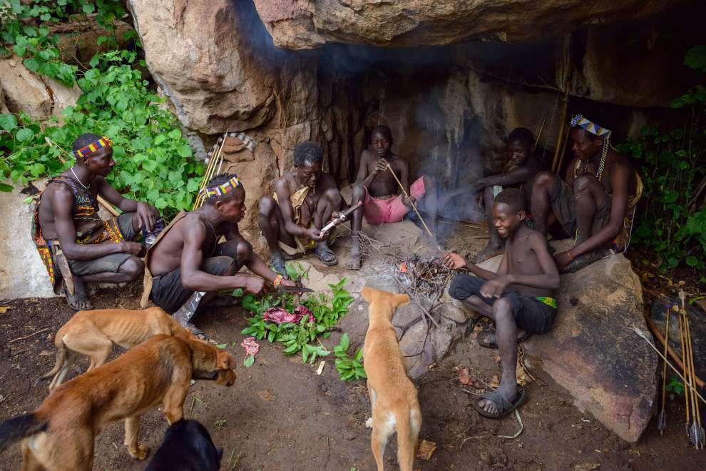 A group of Hadzabe men and boys wearing traditional animal furs sitting around a small campfire inside a shallow rock cave shelter, with hunting dogs standing nearby.