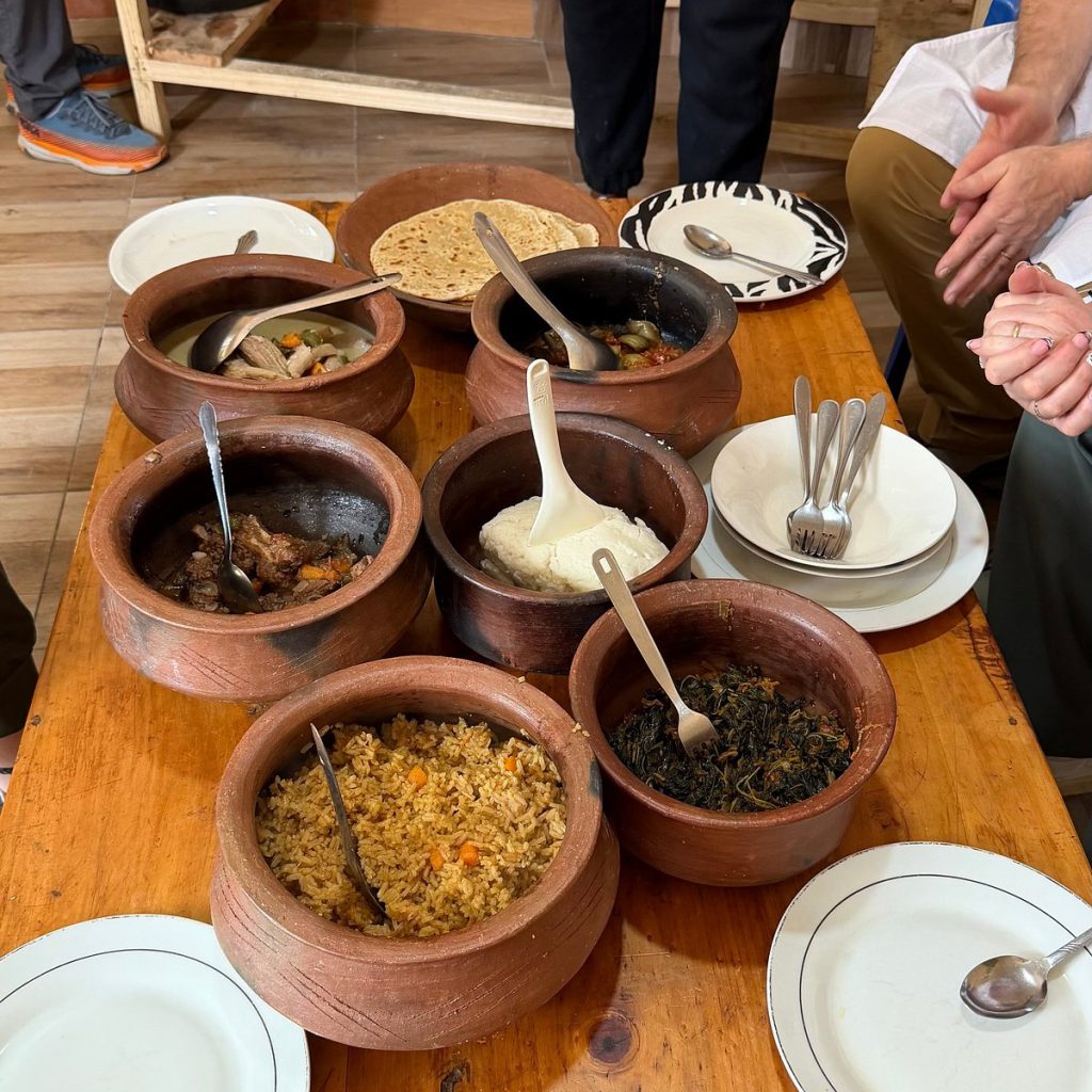 A rich spread of traditional Tanzanian dishes served inside rustic clay pots on a smooth wooden dining table, featuring aromatic pilau rice, ugali, stewed meat, vegetables, and flat chapati bread.