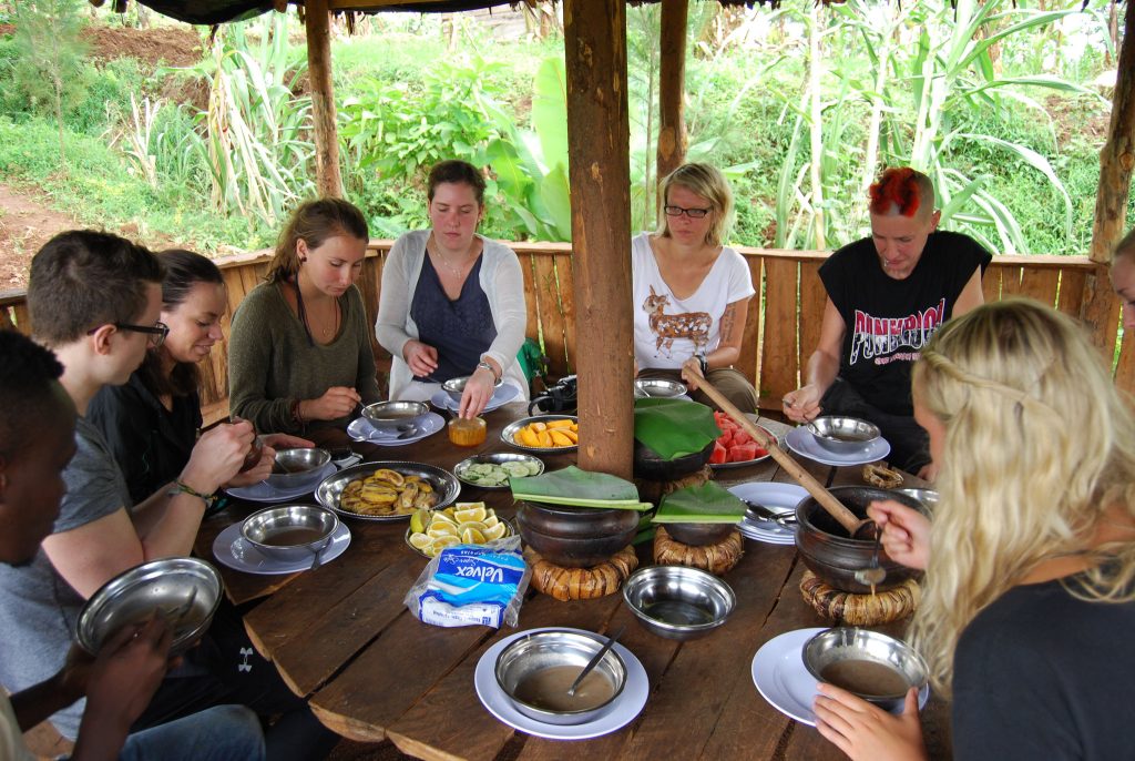 A group of seven travelers sitting around a large, circular wooden table under a thatched roof hut, enjoying a traditional Tanzanian meal served out of rustic clay pots.