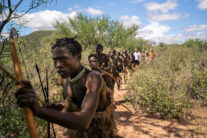 A young Hadzabe hunter leading a group through the bush while holding a wooden bow and wearing a beaded necklace, with other tribesmen and a line of tourists following behind on a dirt path under a cloudy sky.