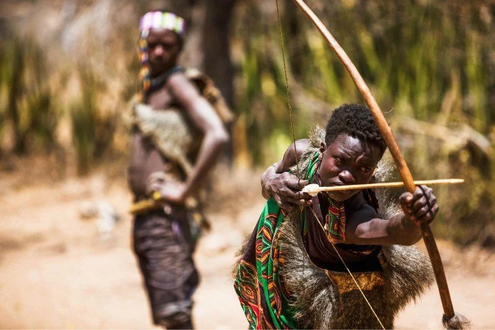 A young Hadzabe hunter focusing intently while pulling back a large wooden bow and arrow, wrapped in a traditional animal fur and patterned green cloth, with another tribesman standing blurred in the background.