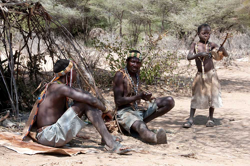 Two Hadzabe men sit on the sandy ground near a brush shelter while talking, as a small boy stands beside them holding a traditional handmade stringed instrument.