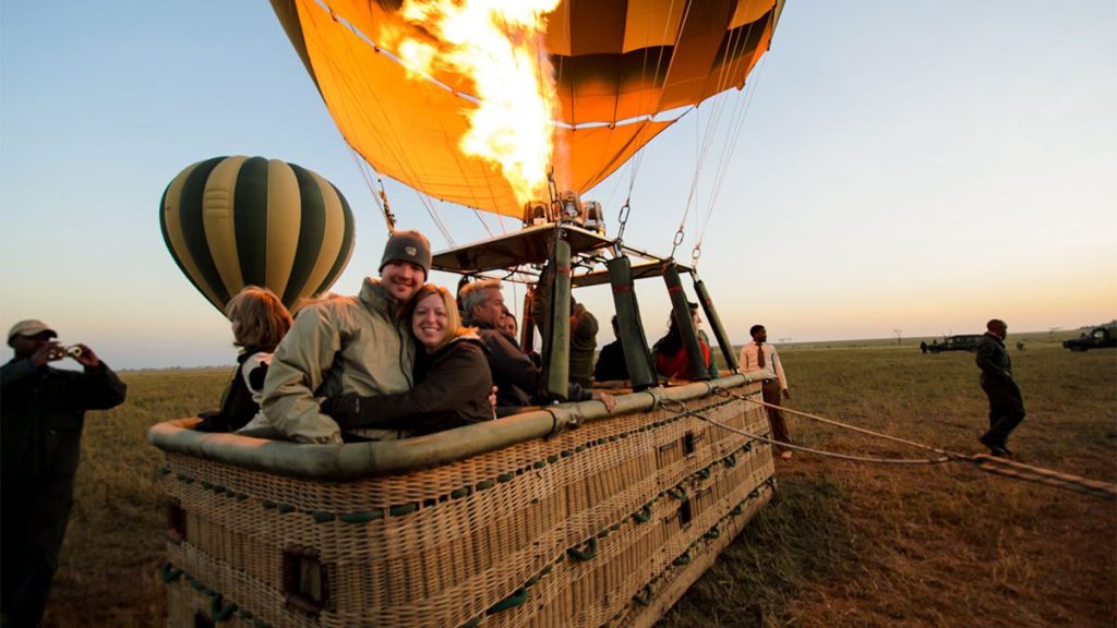 A large wicker basket of a hot air balloon sits on a grassy plain at dawn as a couple smiles for a photo. A bright orange flame erupts from the burner above to inflate the envelope, while another green and gold striped balloon stands inflated nearby in the background.