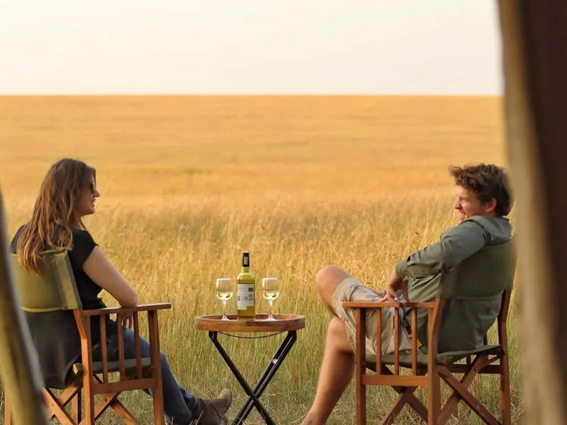 A man and woman sit facing each other in wooden director's chairs on a grassy savanna, smiling over a small table with a bottle of white wine and two filled glasses against an endless golden horizon.