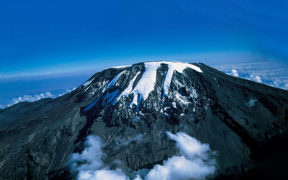 An aerial view of the snow-capped peak of Mount Kilimanjaro, showcasing the glaciers contrasting against dark volcanic rock under a deep blue sky with clouds ringed around its base.