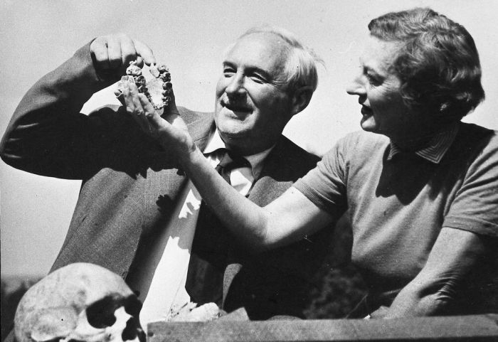 A historical black-and-white photograph showing paleoanthropologists Louis and Mary Leakey sitting together outdoors, closely examining a large fossilized hominin jawbone fragment.