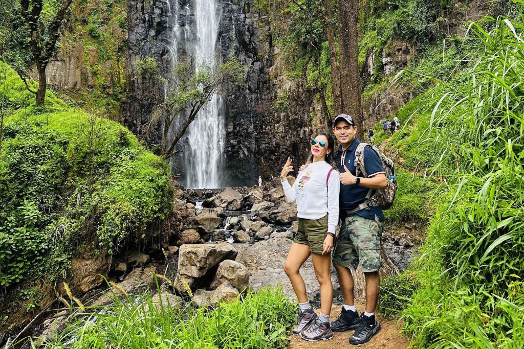 A man and a woman wearing casual hiking clothes and sunglasses posing together on a dirt trail directly in front of the rushing Materuni Waterfall and its rocky base.