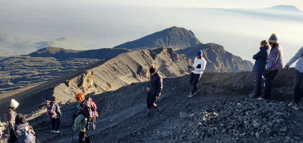 A group of hikers in cold-weather gear walking along a steep, rocky volcanic crater rim with a vast mountain landscape and hazy horizon in the background.