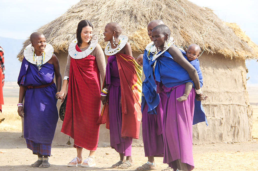 A female tourist wearing a red shúkà wrap and beaded collar walks and laughs alongside several Maasai women carrying young children outside a traditional mud-and-thatch boma hut.
