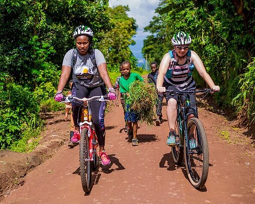 Two female cyclists wearing helmets ride mountain bikes down a red dirt road flanked by dense green foliage, while local children walk behind them carrying bundles of grass.
