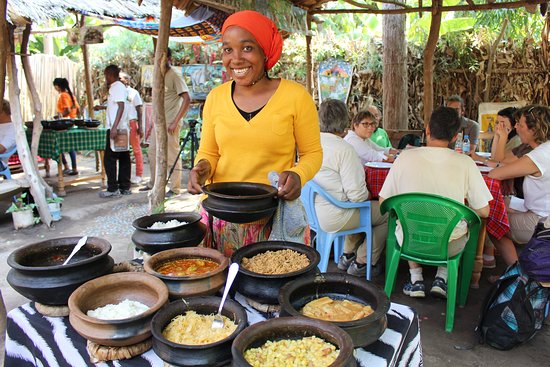 Tanzanian woman wearing a bright yellow shirt and an orange headwrap stands behind a table serving a buffet of traditional local dishes from black clay pots to seated international tourists under a thatched roof.