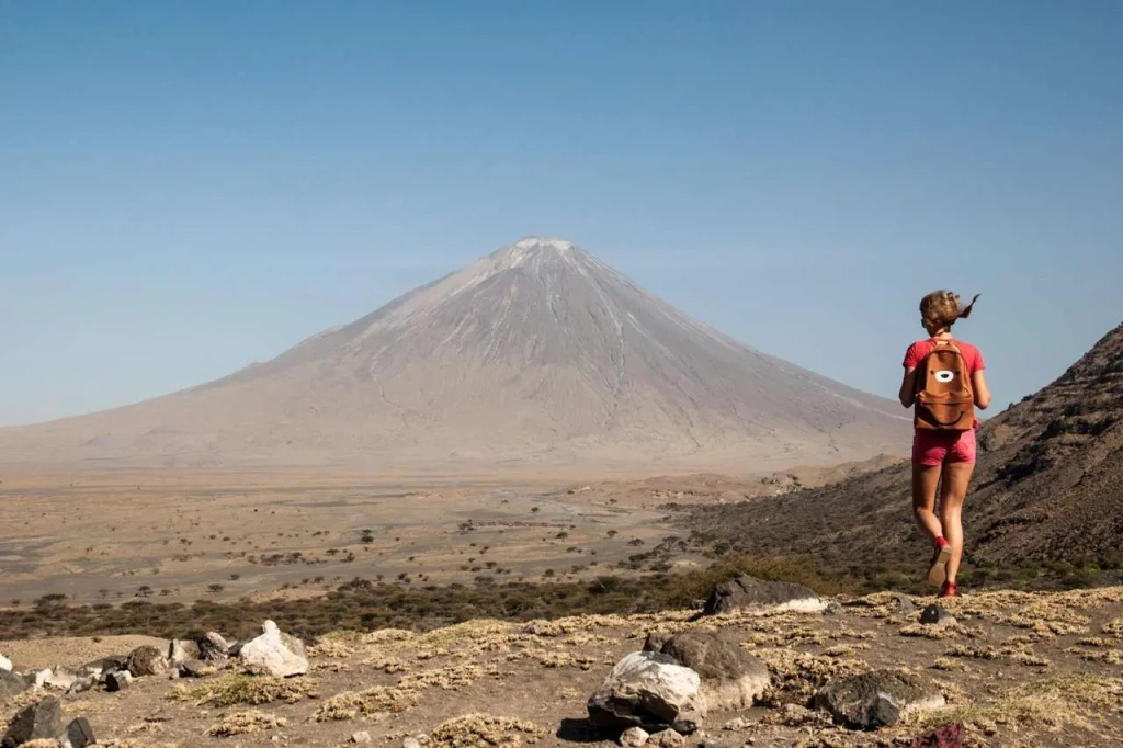 A distinct view of Ol Doinyo Lengai highlighting its asymmetric crater structure and the sheer scale of the mountain. While the lower sections feature patches of hardy volcanic vegetation, the barren, calcified rim serves as a dramatic campsite or resting area for researchers and seasoned trekkers checking the activity of the volcano's inner core.