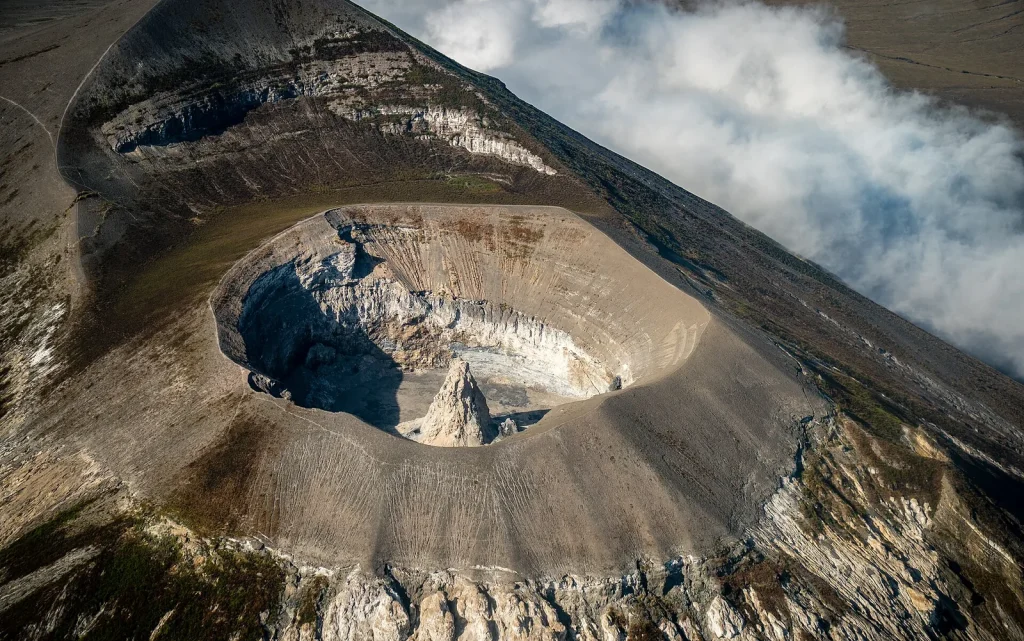 high-angle aerial view looking into a deep, bowl-shaped volcanic crater pit with a sharp, pointed light grey hornito spire rising directly from its center, flanked by steep volcanic slopes and white clouds.