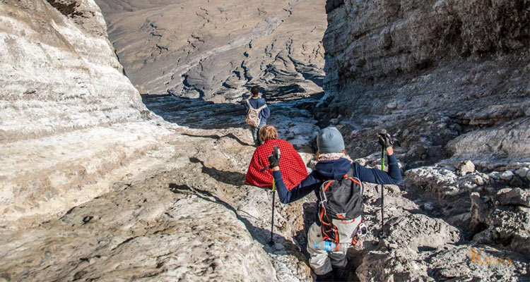Three hikers wearing backpacks and using trekking poles walk carefully down a steep, rugged, light grey volcanic rock and ash canyon trail winding between two layered rock walls.