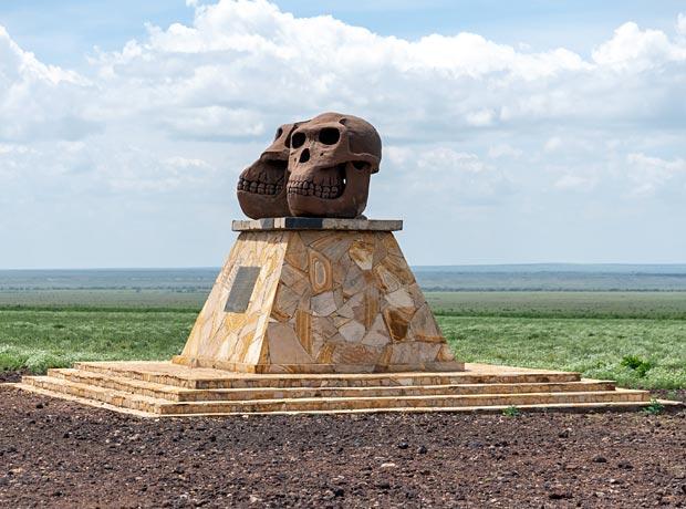 A stone monument featuring two large replica hominin skulls sitting atop a tiered, stone-mosaic pedestal overlooking the expansive, flat grassy plains of Olduvai Gorge.