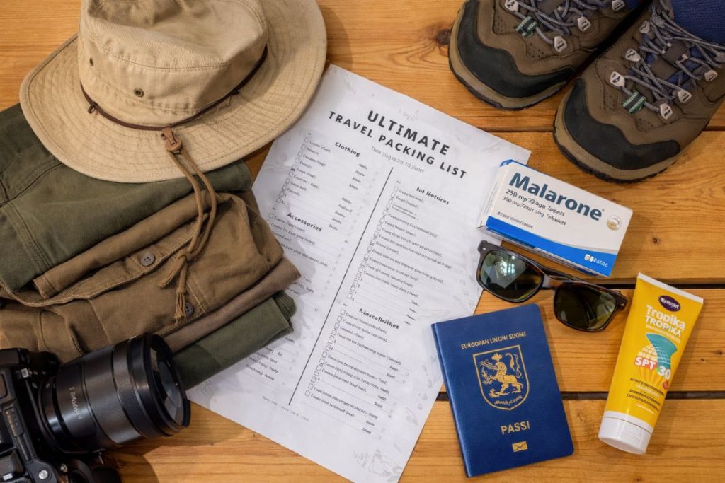 Flat lay of safari travel essentials laid out on a light wooden table, including a wide-brimmed khaki hat, green cargo pants, a camera lens, a printed packing list, a Finnish passport, sunglasses, sunscreen, hiking boots, and a box of Malarone antimalarial medication.