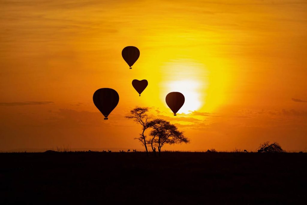 Four black silhouettes of hot air balloons floating in the sky during a bright orange and yellow sunrise over a dark, flat savannah with trees.