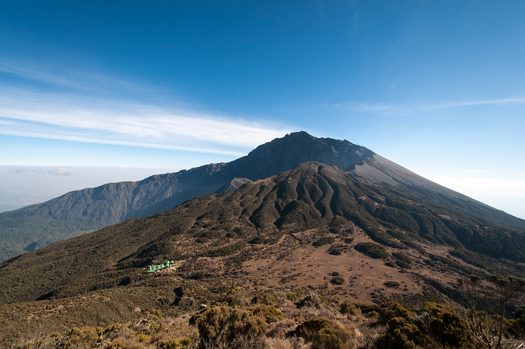 A wide view from high above looking down at a large, grey volcanic ash cone nestled inside a massive volcanic crater, with steep, jagged rocky mountain walls rising high under a bright blue sky and white clouds drifting below.