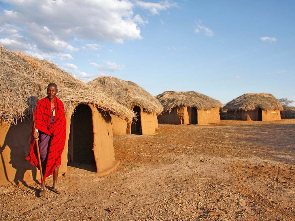 A Maasai man in a red and black plaid wrap stands with a wooden walking stick outside a row of low, rounded mud houses with thick straw and grass roofs.